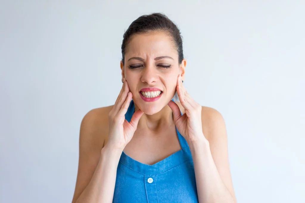 Annoyed young woman suffering from bruxism and touching jaw. Irritated girl with closed eyes gritting teeth from pain.